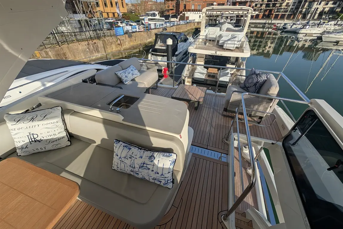 Luxury yacht deck featuring cushioned seating, a wooden table, and decorative cushions by the water, with boats in the background.